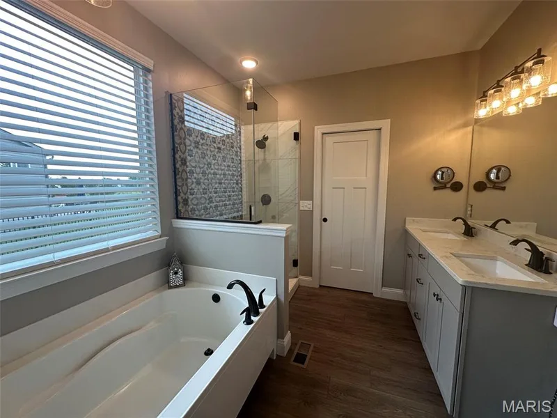 Bathroom featuring a built-in tub with a black faucet, a walk-in shower with glass enclosure, a dual vanity with light-colored countertops, and wood-finish flooring — 110 Alta Mira Court, Dardenne Prairie, MO 63368