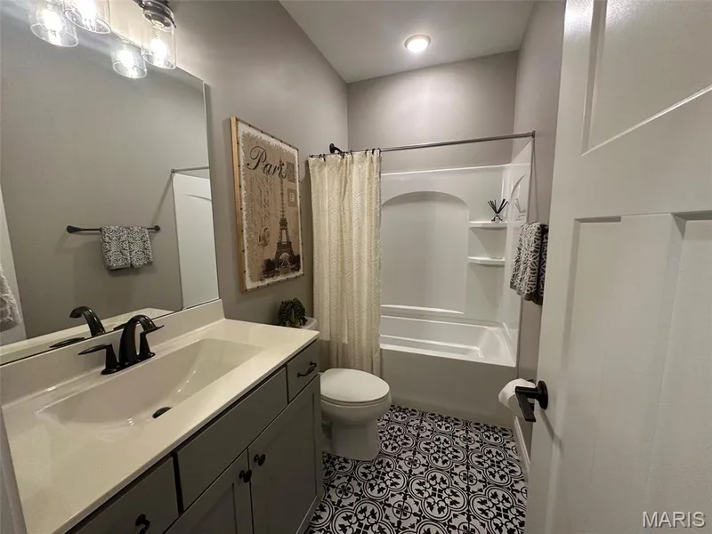 Bathroom featuring black and white patterned floor tiles, a single vanity with a white countertop and dark cabinetry, a combined bathtub and shower unit, a dark-framed wall mirror, and a three-light vanity fixture — 110 Alta Mira Court, Dardenne Prairie, MO 63368