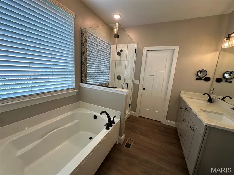 Bathroom featuring a soaking tub with matte black fixtures, a walk-in shower with clear glass enclosure, a dual vanity with light-colored countertops, wood-finish flooring, and a white interior door with dark hardware — 110 Alta Mira Court, Dardenne Prairie, MO 63368