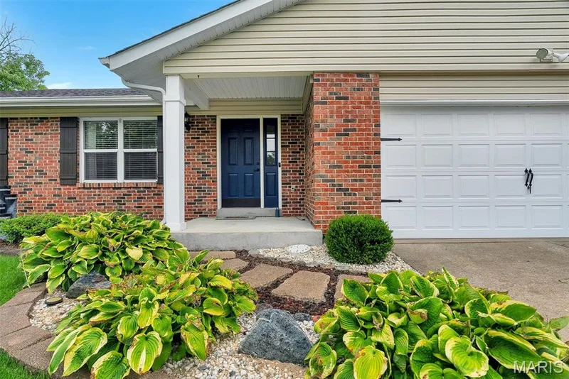 Welcoming front elevation featuring red brick construction, a covered entry portico with white columns, navy blue entry door, and a two-car garage with white panel doors — 104 Cedar Crest Court, St Charles, MO 63304