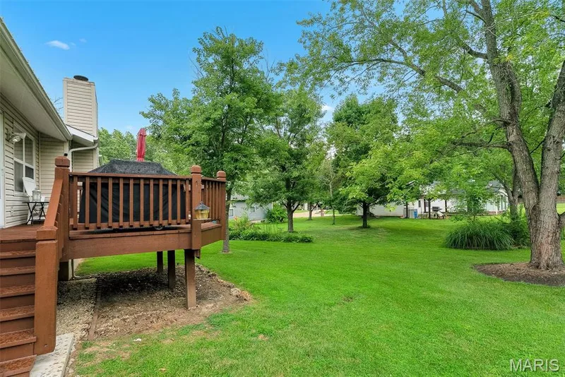 Expansive backyard featuring a manicured lawn, mature shade trees, and an elevated wood-finish deck with railings—ideal for outdoor entertaining and recreation — 104 Cedar Crest Court, St Charles, MO 63304
