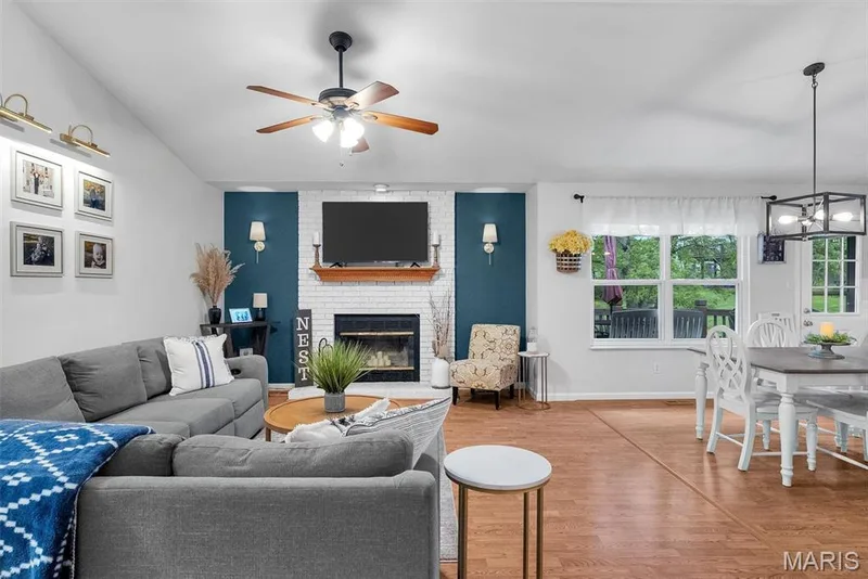 Vaulted ceiling, wood-finish flooring, white brick fireplace with wood mantel, ceiling fan, and modern rectangular chandelier — 104 Cedar Crest Court, St Charles, MO 63304