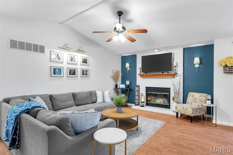 Vaulted ceiling with ceiling fan, white brick fireplace with mantel, two built-in wall sconces, accent wall, and wood-finish flooring — 104 Cedar Crest Court, St Charles, MO 63304