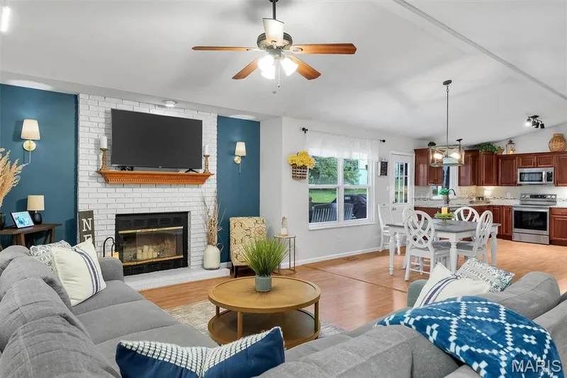 Open-concept living space featuring a fireplace with white brick surround, wood mantel, and sconce lighting — 104 Cedar Crest Court, St Charles, MO 63304