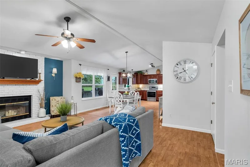 Open-concept living space featuring wood-finish flooring, a white brick fireplace with a wood mantle, and a ceiling fan — 104 Cedar Crest Court, St Charles, MO 63304