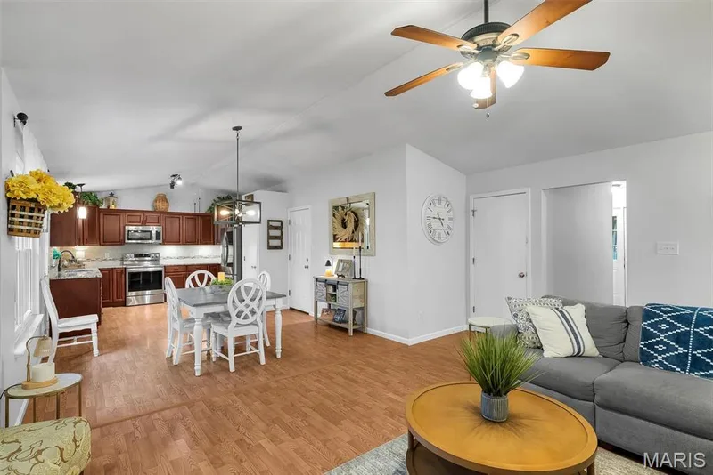 Open-concept living area featuring wood-finish flooring throughout, a fully equipped kitchen with stainless steel appliances and wood cabinetry, a ceiling fan with integrated lighting, and a comfortable seating area with a round wood table — 104 Cedar Crest Court, St Charles, MO 63304
