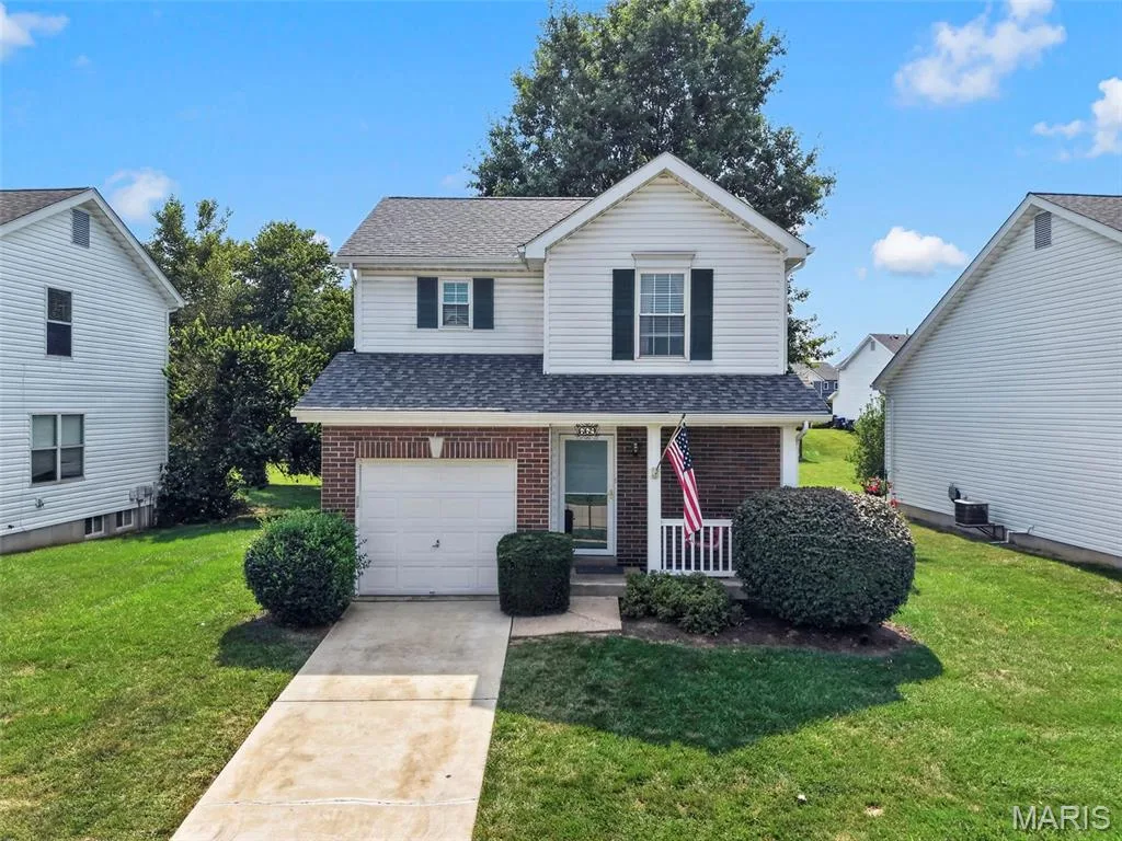 Charming two-story colonial with brick and vinyl siding, featuring a covered front porch with white railings, attached single-car garage, and manicured landscaping with mature trees and established shrubs throughout the property — 669 Homerun Drive, O'Fallon, MO 63366