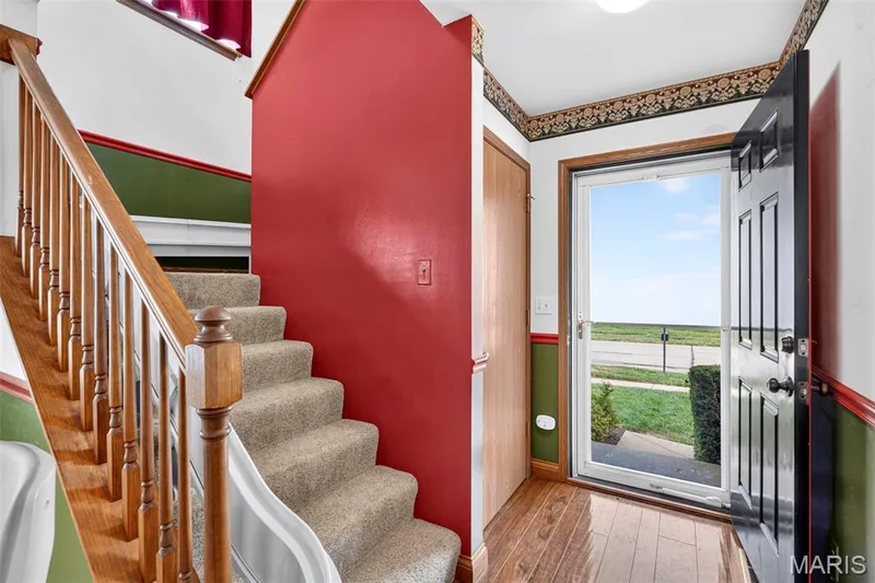 Wood-finish flooring at entry, carpeted staircase, wood banister with balusters, and a solid wood door — 669 Homerun Drive, O'Fallon, MO 63366
