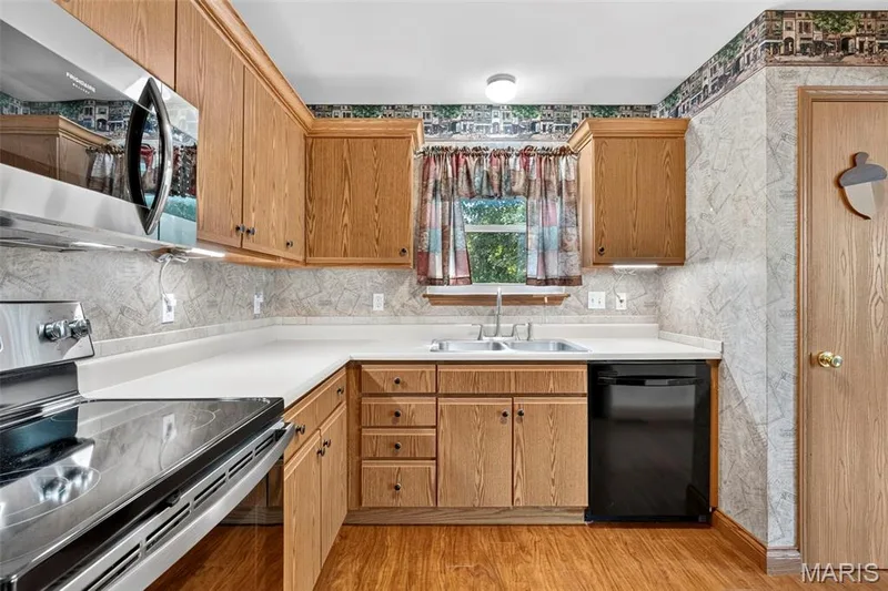 Efficient kitchen featuring wood cabinetry with ample storage, stainless steel appliances including electric range and dishwasher, laminate countertops, ceramic tile backsplash, a window over the sink, and wood-finish flooring throughout — 669 Homerun Drive, O'Fallon, MO 63366