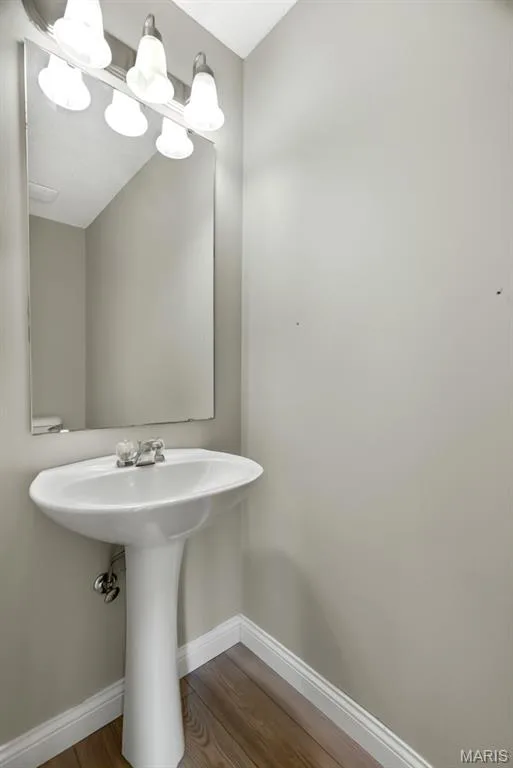 Main Floor Bath. Pedestal sink with chrome fixtures, wood-finish flooring, wall-mounted mirror, and a four-light vanity fixture — 14 Bridgeton Court, Wentzville, MO 63385