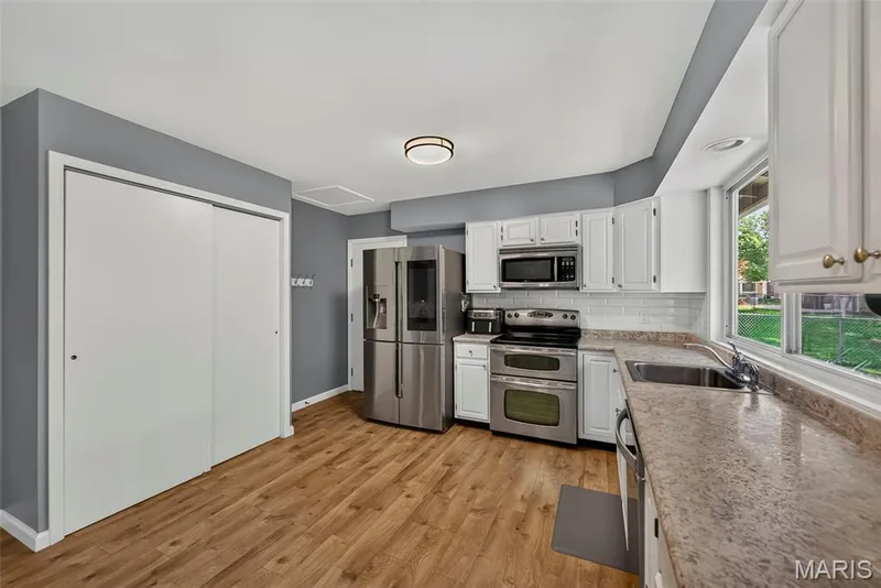Modern kitchen featuring stainless steel appliances, including a double oven and refrigerator with French doors, white cabinetry with ample storage, and laminate wood-finish flooring throughout — 23 Crescent Woods Drive, St Peters, MO 63376
