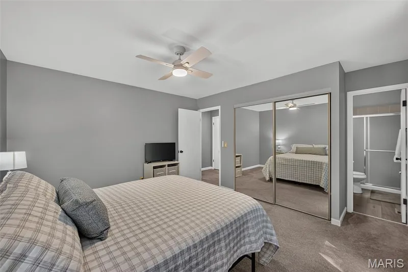 Carpeted bedroom featuring neutral gray walls, a ceiling fan, mirrored closet doors, and an en-suite bathroom with a glass-enclosed shower and toilet — 23 Crescent Woods Drive, St Peters, MO 63376