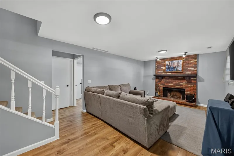 Spacious open-concept recreation room featuring laminate flooring, recessed ceiling lighting, and a built-in brick fireplace with wooden mantelpiece — 23 Crescent Woods Drive, St Peters, MO 63376