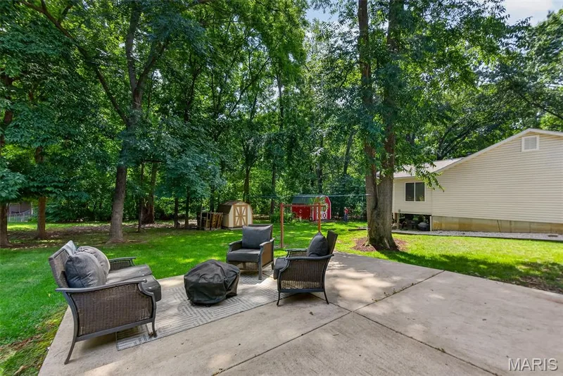 Expansive outdoor living space featuring a concrete patio surrounded by mature shade trees and manicured lawn — 23 Crescent Woods Drive, St Peters, MO 63376