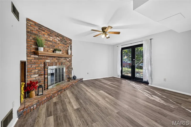 Spacious living area featuring wide-plank wood-finish flooring, exposed brick fireplace with built-in shelving and mantel display, ceiling fan with brass finish, and glass French doors opening to outdoor space — 1319 Brandywine Lane, St Peters, MO 63376