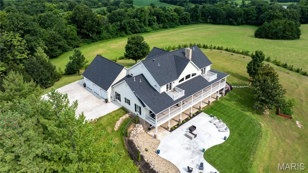 Gable roofline residence with white siding, a three-car garage, expansive covered porch, and a stone chimney — 205 Howell Road, Defiance, MO 63341