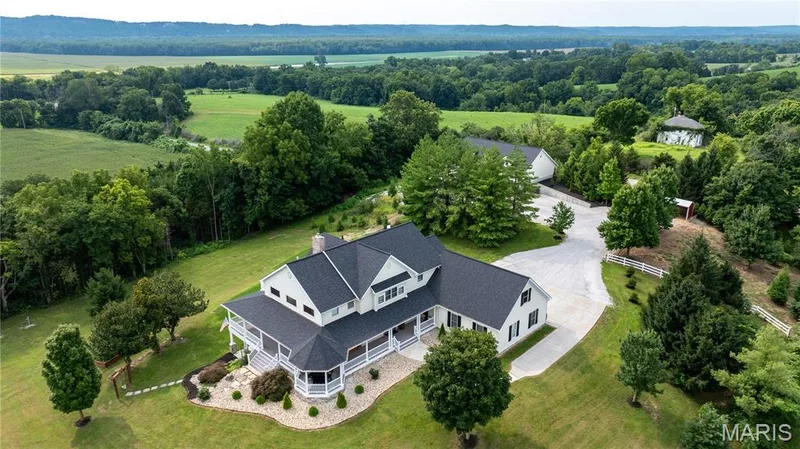 White exterior with dark shutters, dark roof, wraparound porch, and extensive green landscaping — 205 Howell Road, Defiance, MO 63341