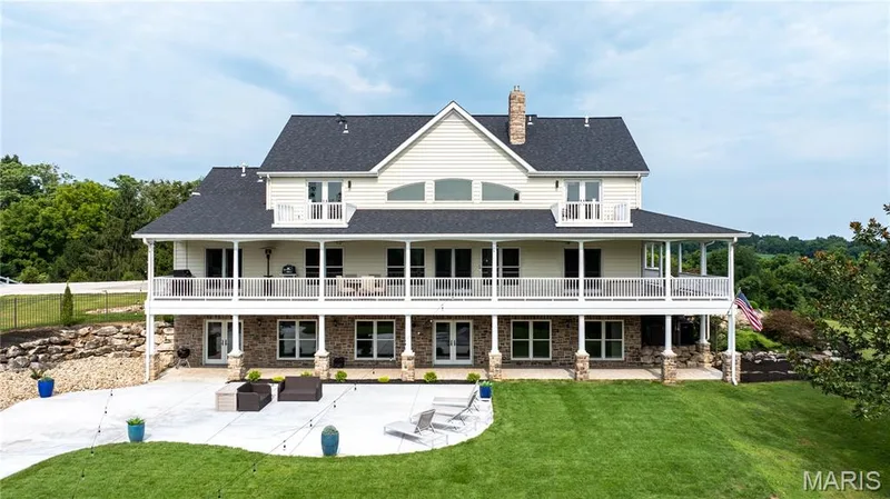 Expansive rear elevation featuring a full-width covered porch, multiple balconies, a large stone patio, and a manicured lawn — 205 Howell Road, Defiance, MO 63341