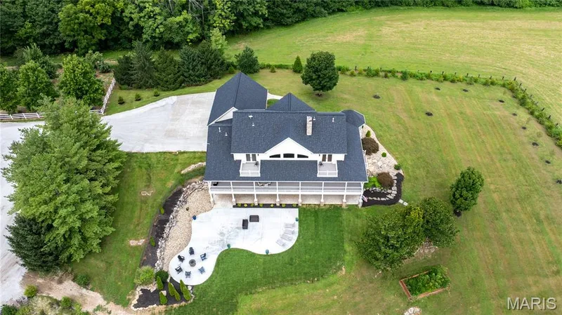 Expansive property featuring a large stone patio, a multi-level deck, dark shingle roofing, mature trees, and a spacious green lawn — 205 Howell Road, Defiance, MO 63341