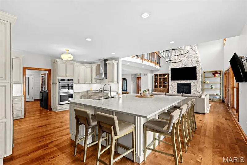Spacious open-concept interior featuring light wood-finish flooring, a contemporary kitchen island with seating, integrated double ovens, a stone-clad fireplace, and an upper-level balcony — 205 Howell Road, Defiance, MO 63341