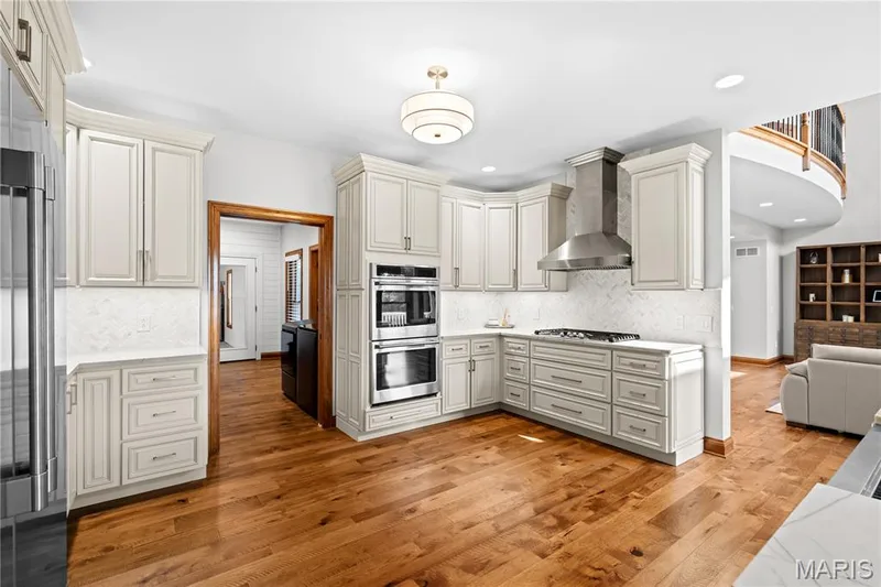Gourmet kitchen featuring hardwood flooring, light-toned cabinetry, stainless steel appliances, a range hood, and a decorative tile backsplash — 205 Howell Road, Defiance, MO 63341