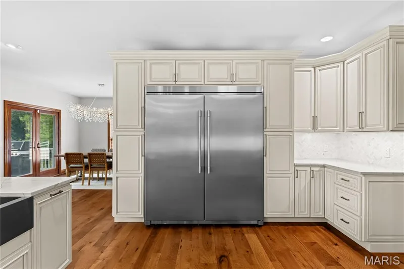 Expansive kitchen featuring wood-finish flooring, custom cabinetry with crown molding, stainless steel refrigerator, white countertops, and a white tile backsplash — 205 Howell Road, Defiance, MO 63341