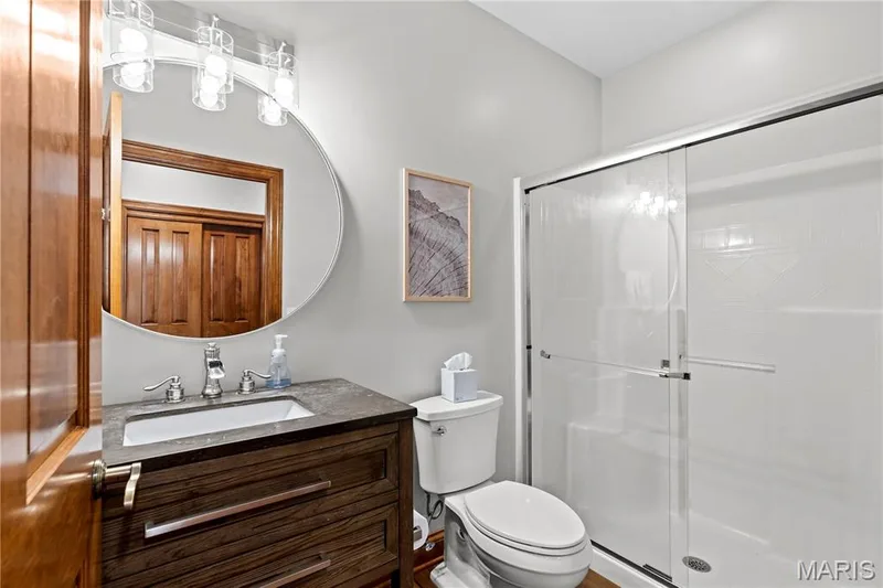 Vanity with integrated sink and stone-finish countertop, clear glass shower enclosure, round wall-mounted mirror, and wall-mounted light fixture — 205 Howell Road, Defiance, MO 63341