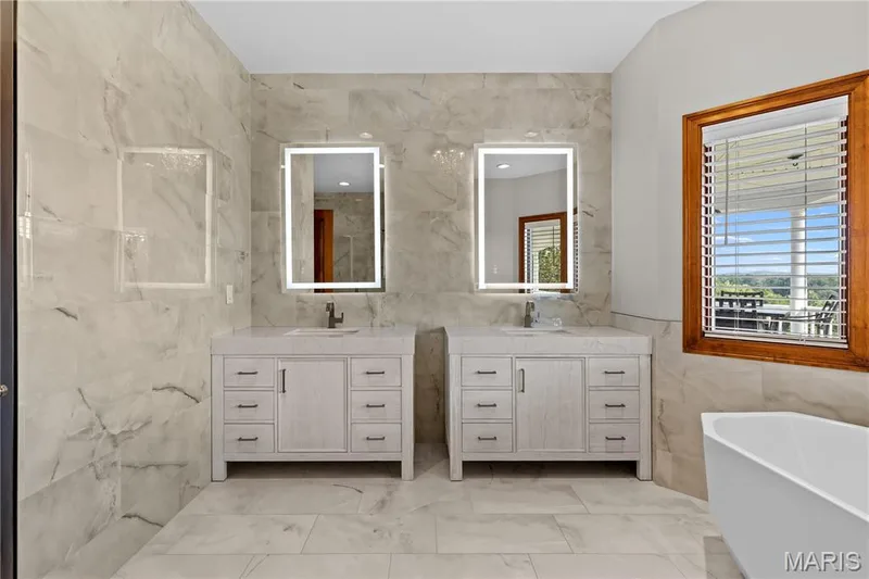 Dual light-up vanity mirrors, two single vanities with integrated sinks, wood-finish window frame, and a freestanding soaking tub — 205 Howell Road, Defiance, MO 63341
