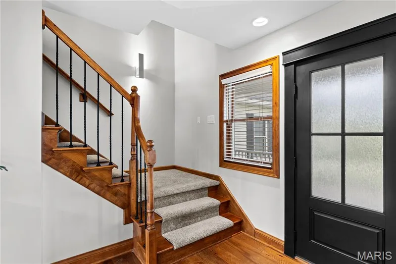 Hardwood flooring with carpeted stairs, a dark wood-framed window with blinds, and a black paneled door with frosted glass inserts — 205 Howell Road, Defiance, MO 63341