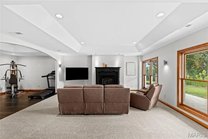 Recessed lighting, tray ceiling, fireplace with detailed mantel, large windows with wood trim, and a separate arched alcove with dark wood-finish flooring — 205 Howell Road, Defiance, MO 63341