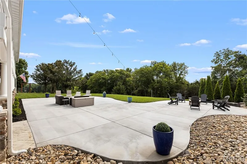 Expansive concrete patio with decorative string lighting, surrounded by a manicured lawn and mature trees — 205 Howell Road, Defiance, MO 63341
