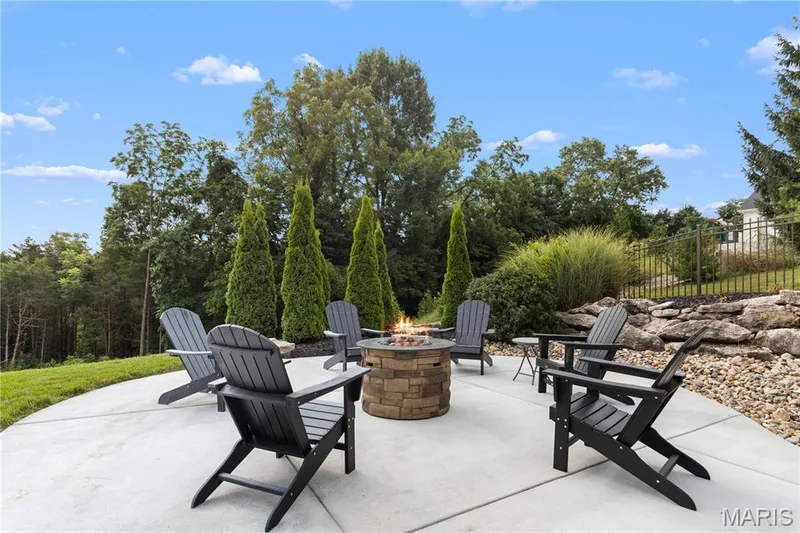Expansive concrete patio with a built-in stone fire pit, surrounded by mature trees and landscaped rock retaining walls — 205 Howell Road, Defiance, MO 63341