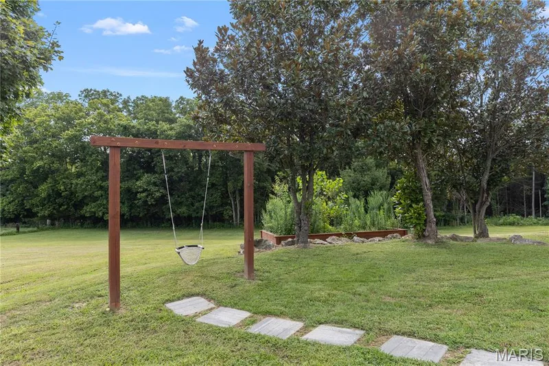 Expansive green space featuring a wooden swing set, stepping stone path, mature trees, and a raised garden bed — 205 Howell Road, Defiance, MO 63341