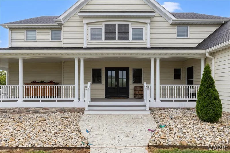 Exterior featuring light-toned siding, a covered porch with white columns and railings, and a stamped concrete pathway — 205 Howell Road, Defiance, MO 63341