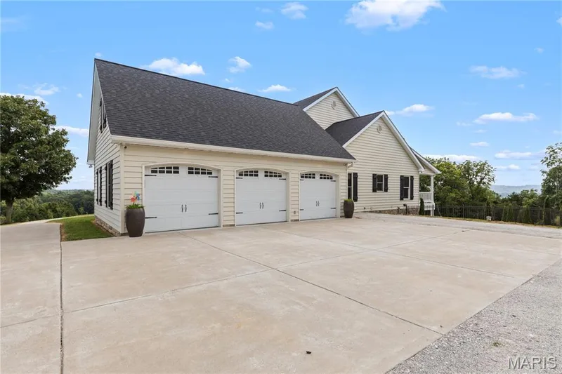 Exterior featuring light-colored siding, a dark shingle roof, three garage bays with white doors, and black window shutters — 205 Howell Road, Defiance, MO 63341