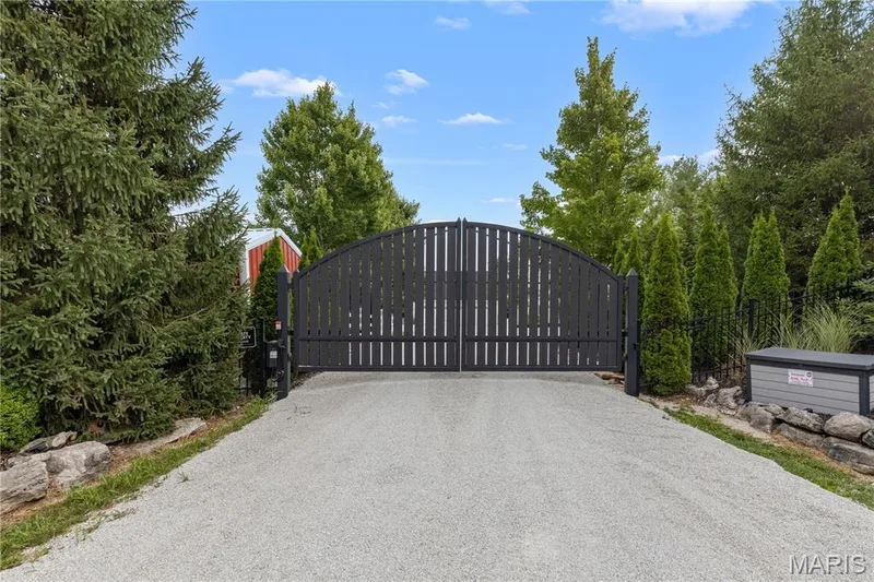Gravel driveway with an arched privacy gate, bordered by mature trees and landscaping — 205 Howell Road, Defiance, MO 63341