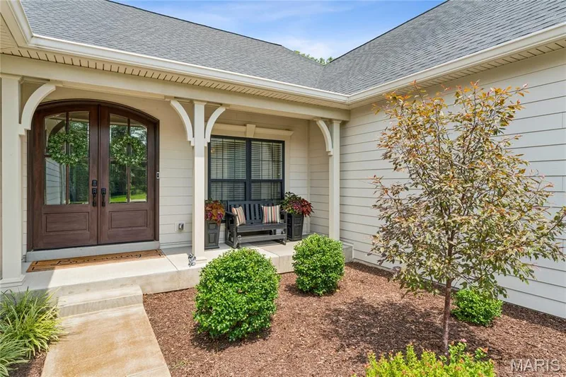 Arched double entry doors, covered front porch, horizontal siding exterior, and a manicured landscape with mature plantings — 5 Timberdel Place, Foristell, MO 63348