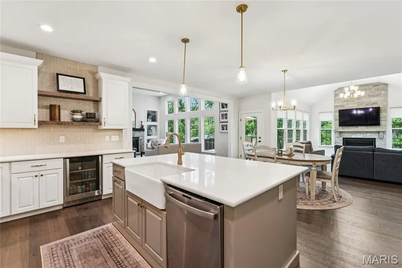 Spacious kitchen featuring an expansive island with a farmhouse sink, stainless steel dishwasher, white cabinetry, and floating wood shelving — 5 Timberdel Place, Foristell, MO 63348