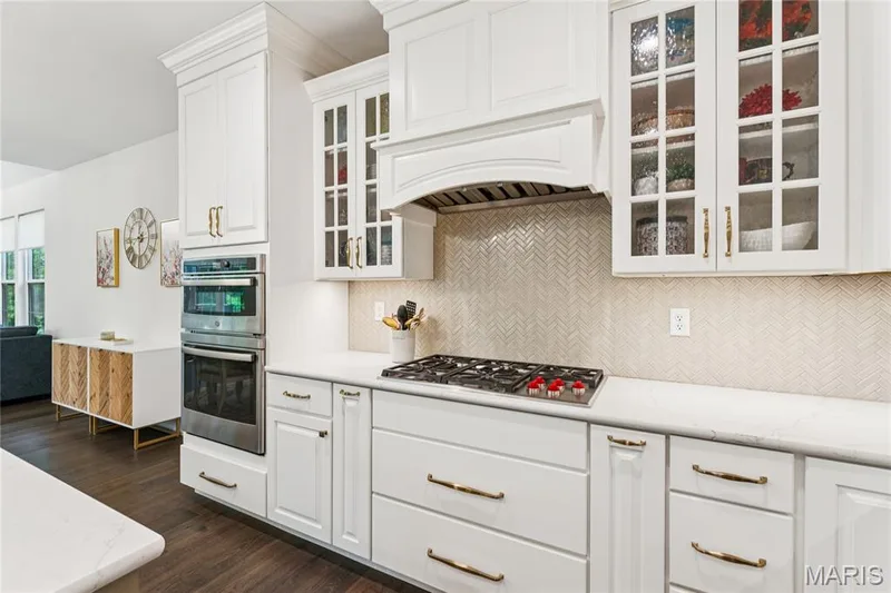 White cabinetry with crown molding, integrated double ovens, a gas cooktop, glass-front upper cabinets, and a herringbone pattern backsplash — 5 Timberdel Place, Foristell, MO 63348