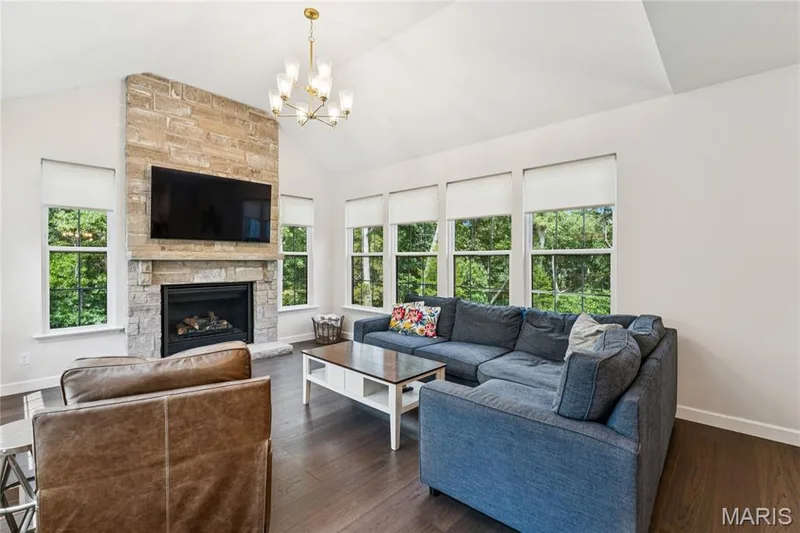 Vaulted ceiling living area featuring a stone-clad fireplace and hearth, wood-finish flooring, and multiple window units — 5 Timberdel Place, Foristell, MO 63348