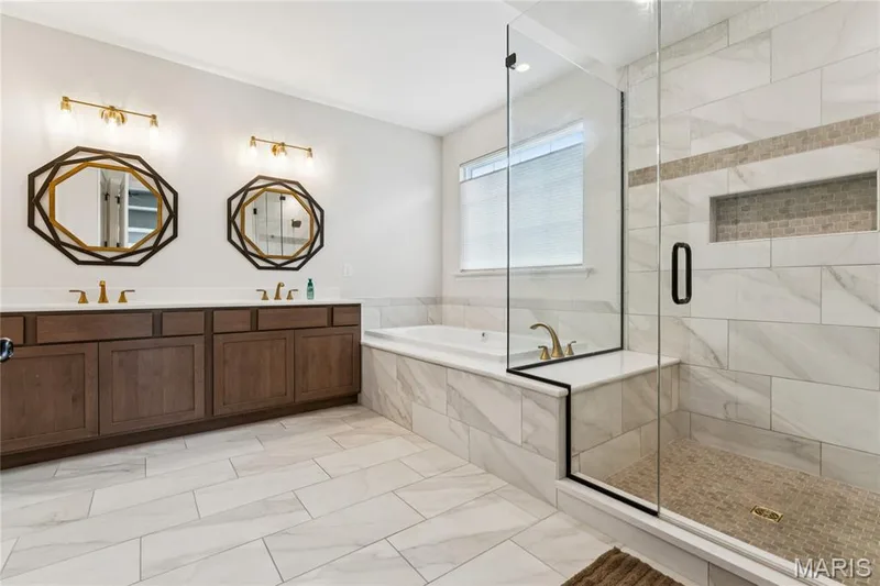 Dual vanity with individual octagonal mirrors, walk-in seated double faucet shower with glass enclosure, built-in bathtub, and large format tile flooring — 5 Timberdel Place, Foristell, MO 63348