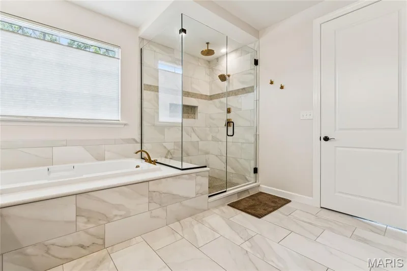 Spacious bathroom featuring a built-in soaking tub, glass-enclosed seated shower with a niche, 2 wall-mounted shower heads, and tile flooring — 5 Timberdel Place, Foristell, MO 63348