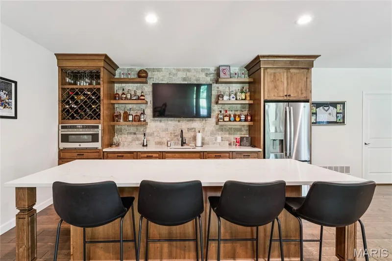 Built-in bar featuring wood cabinetry, stainless steel appliances, a stone tile backsplash, and a large central island with quartz countertop — 5 Timberdel Place, Foristell, MO 63348