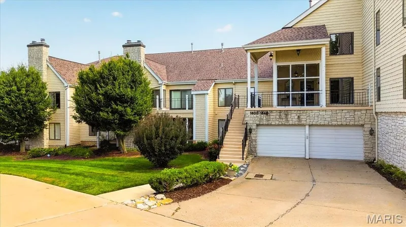 Expansive exterior featuring light-colored siding, a covered balcony with black railing, two visible attached garages, two prominent brick chimneys, and mature landscaping — 1608 Lake Knoll Drive #1A, Lake St. Louis, MO 63367