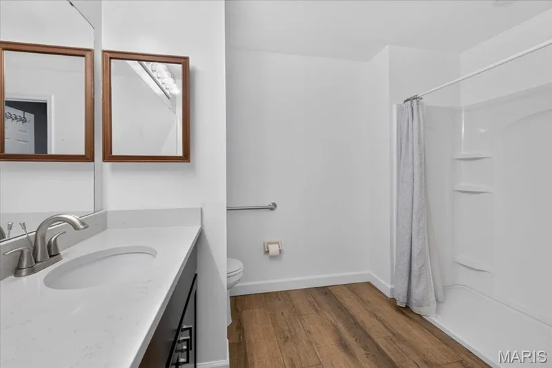 White vanity with an integrated sink, brushed nickel faucet, wood-finish flooring, and a shower with built-in shelving — 1608 Lake Knoll Drive #1A, Lake St. Louis, MO 63367