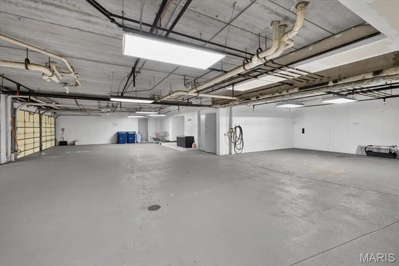Expansive garage with a painted concrete floor, exposed utility pipes, fluorescent lighting, and a roll-up garage door — 1608 Lake Knoll Drive #1A, Lake St. Louis, MO 63367