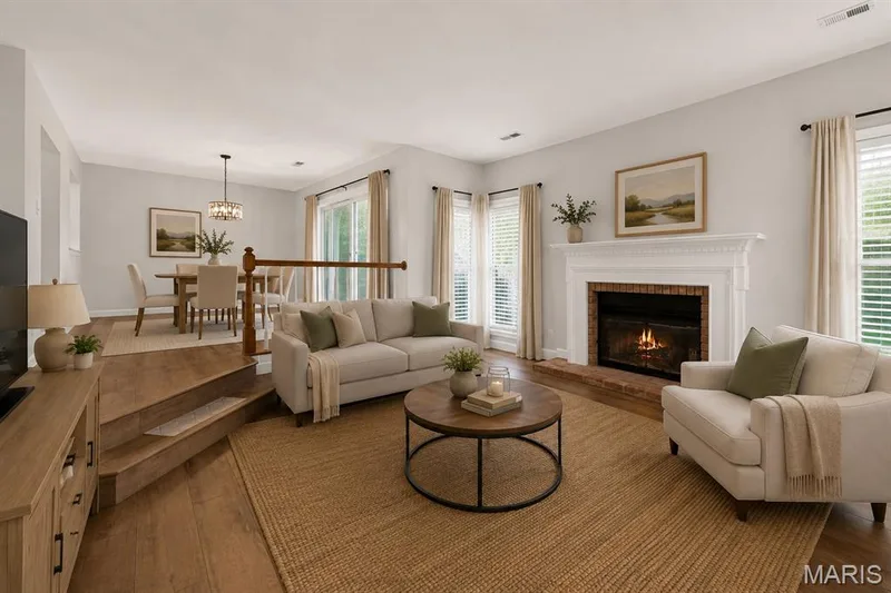 Wood-finish flooring throughout, featuring a brick-surround fireplace with a white mantel, and multiple windows with blinds. Digitally enhanced and virtually staged. — 1608 Lake Knoll Drive #1A, Lake St. Louis, MO 63367