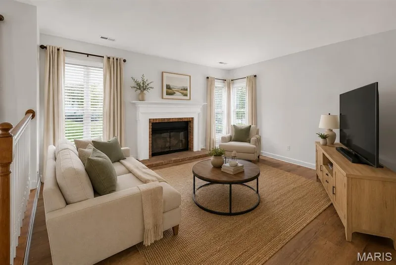 Spacious living area featuring a brick fireplace with a white mantel, wood-finish flooring, and natural light from multiple windows. Digitally enhanced and virtually staged. — 1608 Lake Knoll Drive #1A, Lake St. Louis, MO 63367