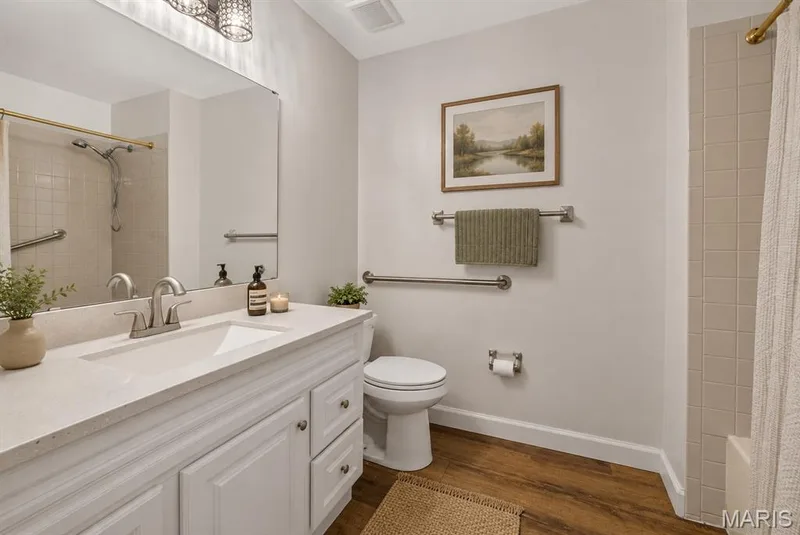 Bathroom featuring a white vanity with an integrated sink, brushed nickel faucet, wood-finish flooring, and a tiled shower with grab bars. Digitally enhanced and virtually staged. — 1608 Lake Knoll Drive #1A, Lake St. Louis, MO 63367