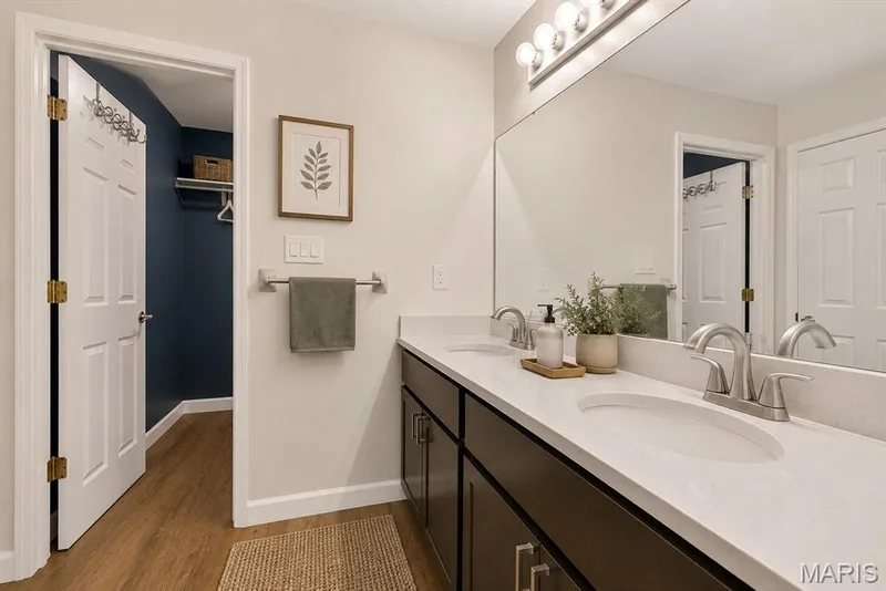 Dual vanity with integrated sinks, white countertop, dark wood-finish cabinetry, and full-width mirror. Digitally enhanced and virtually staged. — 1608 Lake Knoll Drive #1A, Lake St. Louis, MO 63367